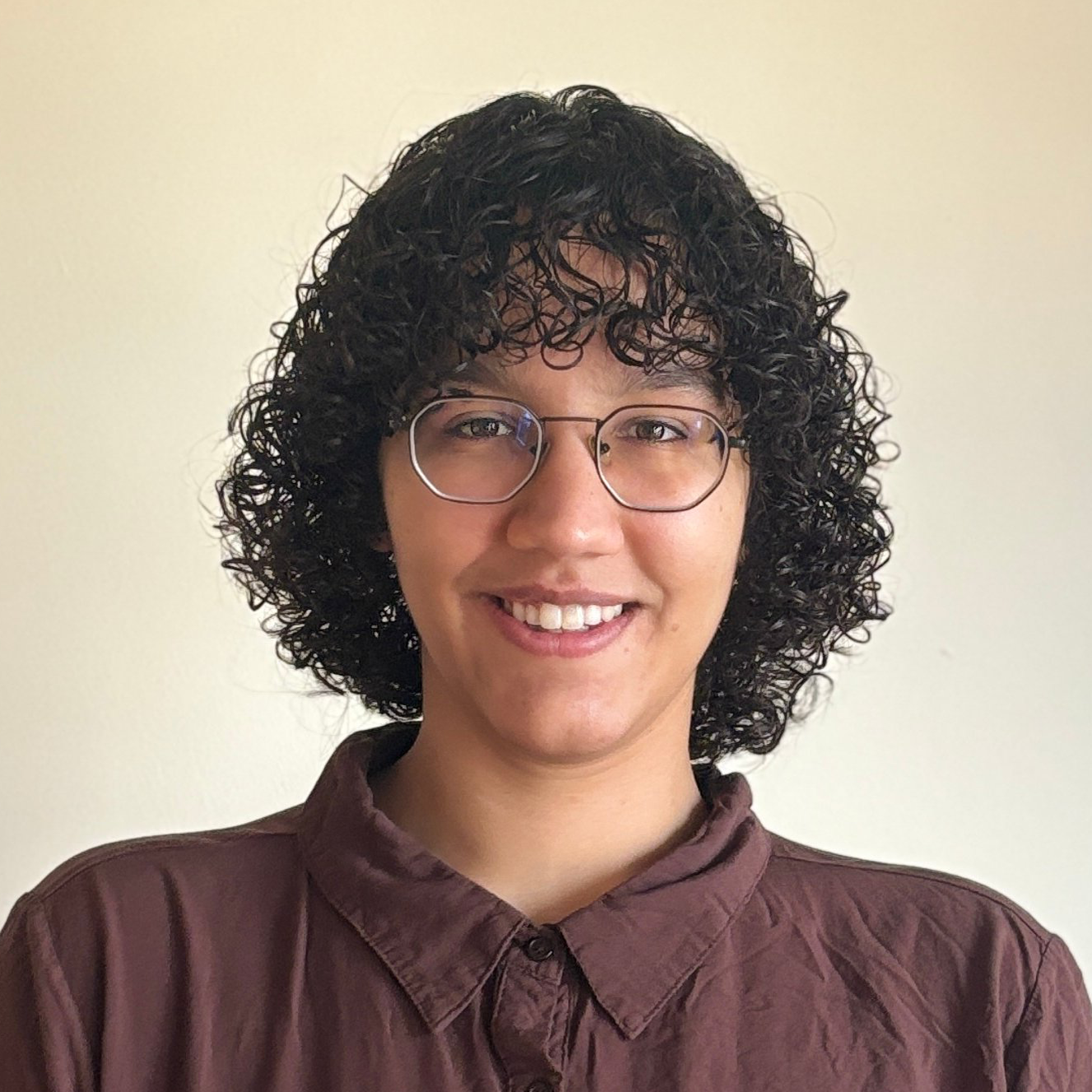 Headshot photo of a person with black glasses, a brown button up shirt, shoulder length curly hair,
                             and brown skin looking straight into the camera and smiling.